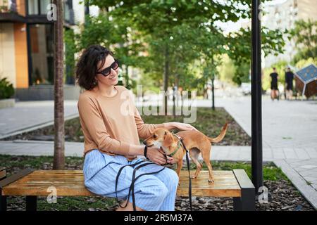 Giovane adulta in città con un cane seduto e abbracciato, sorridente. Accarezzare il cane, nutrirsi, divertirsi Foto di alta qualità Foto Stock