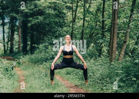 Giovane donna in forma che pratica yoga nella foresta estiva, indossando leggings neri e top Foto Stock