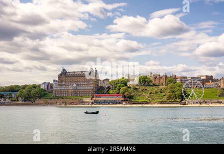 Lungomare di Scarborough con un edificio alberghiero e ruota panoramica sulla spiaggia. Un pescatore in una piccola barca è a metà distanza e un cielo con nuvola è a. Foto Stock