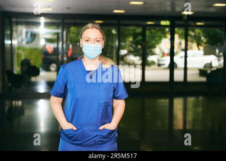 Giovane donna che indossa uniforme blu e maschera facciale, in piedi nel corridoio dell'ospedale Foto Stock