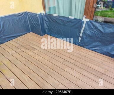 Terrazza in legno marrone preparata per l'oliatura e la pittura con pareti e finestre coperte di film. Pulizia della pavimentazione Foto Stock