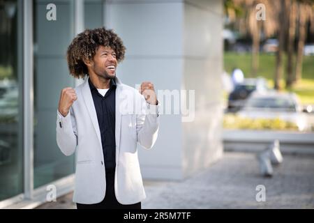 Allegro uomo d'affari nero di mezza età alzando le mani e gioendo alla vittoria vicino alla costruzione di uffici, spazio libero Foto Stock