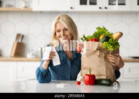 Economia dello shopping. Donna matura in cucina Holding Grocery Bag e conto corrente Foto Stock