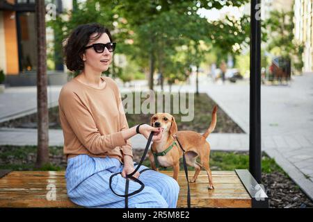 Giovane adulta in città con un cane seduto e abbracciato, sorridente. Accarezzare il cane, nutrirsi, divertirsi Foto di alta qualità Foto Stock