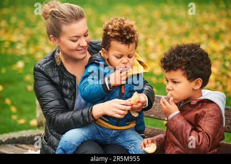 Ritratto all'aperto di felice madre giovane con due figli graziosi, famiglia godendo bella giornata autunnale nel parco pubblico, gara mista famiglia. I bambini mangiano spuntini, re Foto Stock