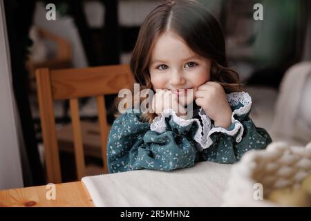 Adorabile bambina con capelli ricci indossa abiti festosi stile rurale è sorridente, guardando la macchina fotografica, seduto accanto al tavolo e sostiene il mento con ha Foto Stock