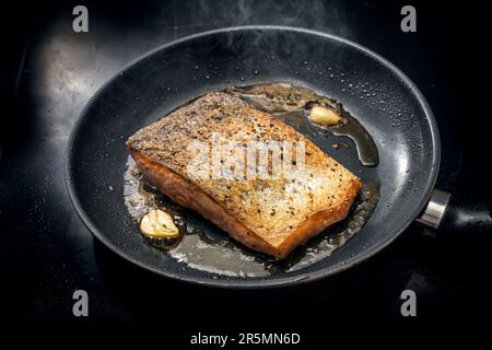 Il filetto di salmone fresco con pelle croccante viene fritto con olio d'oliva, erbe e aglio in una padella nera sul piano cottura, spazio copia, fuoco selezionato, narro Foto Stock