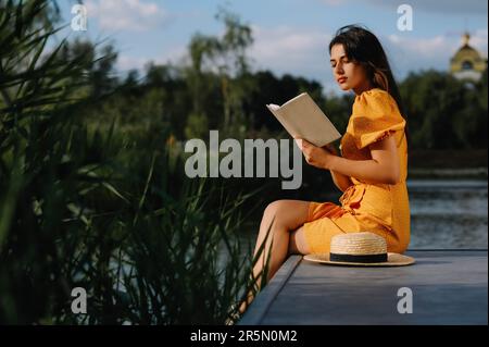 Giovane donna che legge libro vicino al lago nelle giornate di sole Foto Stock