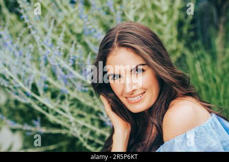 Primo piano esterno ritratto di bella giovane donna con capelli scuri in posa giardino estivo con fiori viola Foto Stock