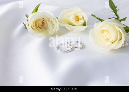 Organizzazione di nozze con due anelli di nozze in platino con diamanti e delicate rose da tè su sfondo bianco satinato. layout per la progettazione Foto Stock