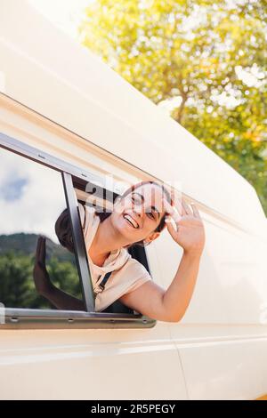 Giovane donna che sventolava attraverso la finestra del suo camper. Viaggio in pulmino e avventura estiva all'aperto. Concetto di stile di vita Nomad Foto Stock