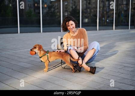 Giovane adulta in città con un cane seduto e abbracciato, sorridente. Accarezzare il cane, nutrirsi, divertirsi Foto di alta qualità Foto Stock