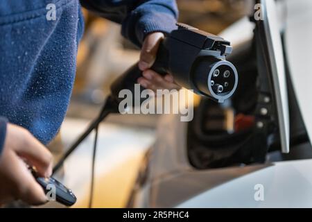 Donna con le mani che reggono il caricabatterie per auto elettrica e il telecomando, pronto per il collegamento al veicolo elettrico nel garage di casa. Foto Stock