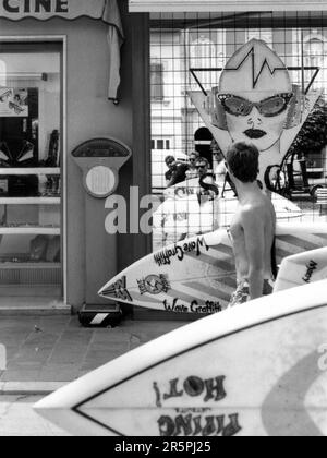 Fotografia di strada in bianco e nero. Tre surfisti con tavole da surf e fotografo si riflettono nel design dello specchio anteriore del negozio mentre camminano. Foto Stock