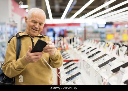 uomo anziano in grigio pensionato esaminano il contatore con gadget elettronici e tablet in showroom del negozio di merci digitali Foto Stock