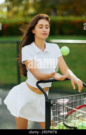 abitudini sane, bella giovane donna con lunghi capelli brunette in piedi in vestito bianco vicino al carrello e lancio di palla in aria, sfondo sfocato, cou tennis Foto Stock