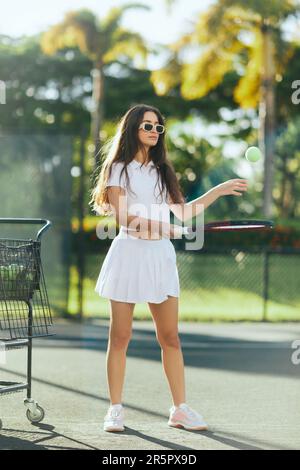 donna bruna elegante e giovane con capelli lunghi in piedi in vestito bianco e occhiali da sole alla moda mentre tiene la racchetta e colpire la palla vicino al golf cart Foto Stock