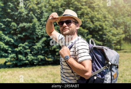 Uomo adulto con occhiali da sole, cappello e zaino che si gode l'estate nel parco della città, guardando la macchina fotografica e sorridendo. Foto Stock