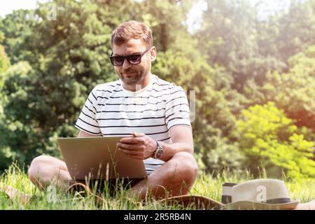 Giovane uomo adulto che indossa occhiali da sole seduti sul prato del parco e utilizza il suo computer portatile. Foto Stock