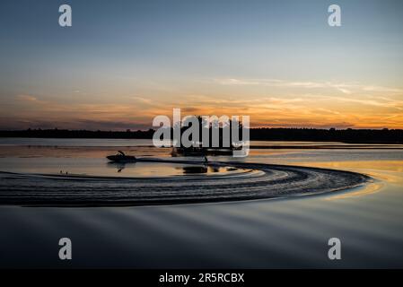 Sci d'acqua wake boarder essere trascinato attraverso il lago calmo da un motoscafo al tramonto con piccola isola sullo sfondo Foto Stock