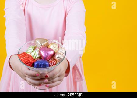 Bambina che tiene la ciotola di vetro con due mani piene di cioccolato avvolto. Idea per banner Sugar Feast. Sfondo giallo, spazio di copia. Foto Stock