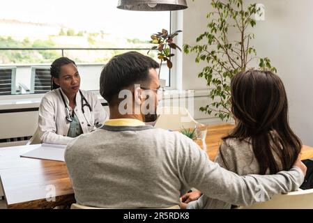 Donna medico visita una giovane coppia sposata nella loro casa e dà le consultazioni sulla salute. Operatrice medica femminile in un appartamento pazienti che ascolta circa Foto Stock