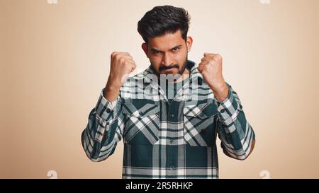 Aggressivo arrabbiato giovane indiano cercando di combattere a macchina fotografica, scuotendo pugno, pugilato con espressione, punizione, disputa. Simpatico ragazzo indù bearded isolato da solo su sfondo beige studio Foto Stock