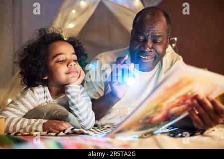 Papà afro-americano che tiene libri e torcia e raccontare storie a suo figlio piccolo mentre giacciono sul pavimento nella stanza Foto Stock