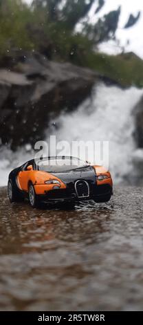 Una piccola macchina giocattolo sportiva parcheggiata in cima ad una grande formazione rocciosa, scoscese, contro una cascata Foto Stock