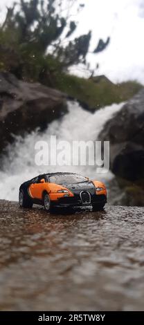 Una piccola macchina giocattolo sportiva parcheggiata in cima ad una grande formazione rocciosa, scoscese, contro una cascata Foto Stock