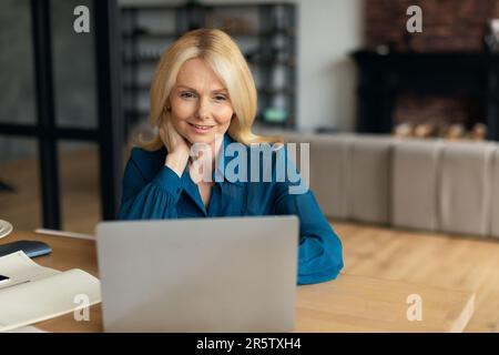 Allegra imprenditrice matura europea che guarda al computer, videochiama nel salotto interno, spazio libero Foto Stock