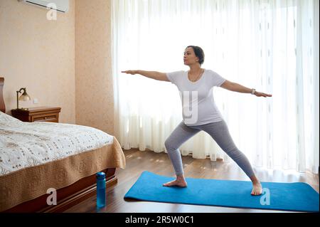 Donna incinta in gravidanza tardiva, pratica yoga a casa, in piedi in guerriero posa sul materassino di esercizio. Verticale a lunghezza intera Foto Stock