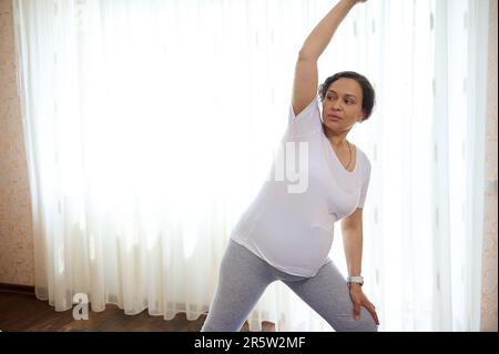 Sicura bella donna incinta multietnica pratica esercizi di stretching prenatale a casa. Sport in gravidanza Foto Stock