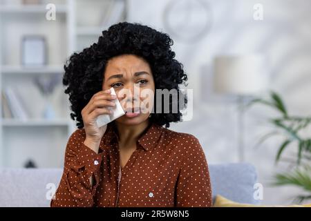Primo piano. Ritratto di giovane afroamericana sconvolta seduta sul divano a casa e piangendo, soffriva di depressione, strappando lacrime con tovagliolo. Foto Stock