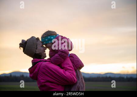 Giovane madre gioiosa che solleva la figlia del bambino in un momento d'amore tra loro fuori al crepuscolo. Foto Stock