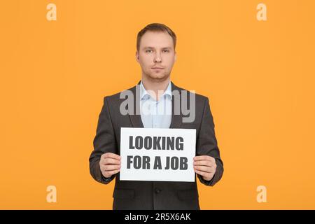 Disoccupato uomo in attesa di segno con frase cercando Un lavoro su sfondo arancione Foto Stock