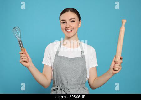 Bella giovane donna in grembiule pulito con utensili da cucina su sfondo azzurro Foto Stock