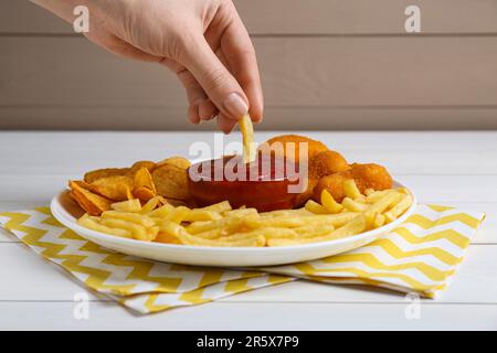 Donna che immerga la frittura francese in ketchup al tavolo di legno bianco, primo piano Foto Stock