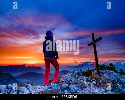 Alpinista accanto alla croce sommitale dell'iceberg e nuvole rosse all'alba, Parco Nazionale Berchtesgaden, Ramsau, Berchtesgadener Land, Alto Foto Stock