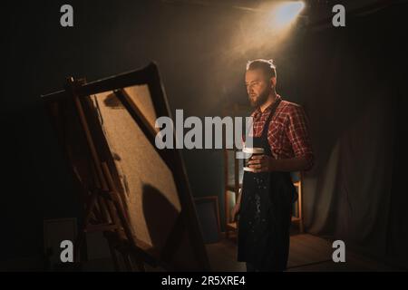 Artista maschile di talento lavora sulla pittura astratta a olio, crea un capolavoro moderno in uno studio creativo scuro con grande tela. Foto Stock