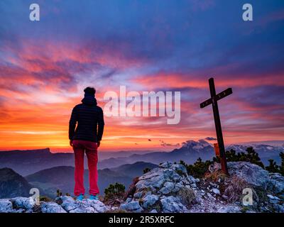 Alpinista accanto alla croce sommitale dell'iceberg e nuvole rosse all'alba, Parco Nazionale di Berchtesgaden, Ramsau, Berchtesgadener Land, alta Baviera Foto Stock