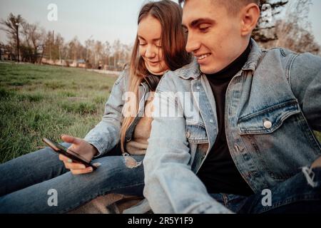 Una giovane coppia alla moda sorride mentre guarda il telefono nel parco Foto Stock