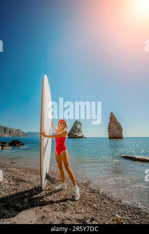 Donna surfista durante il viaggio estivo per le vacanze in spiaggia. Donna di mezza età in costume da bagno rosso e berretto con linfa sullo sfondo del mare e delle rocce Foto Stock