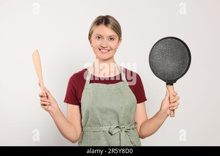 Bella giovane donna in grembiule pulito con utensili da cucina su sfondo grigio chiaro Foto Stock