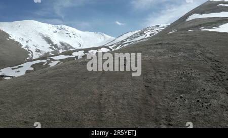 Un giovane si trova sulle alte montagne, coperto di erba verde e di sciagli innevati, una cima innevata di una scogliera. In una giornata di sole contro un cielo blu con Whi Foto Stock