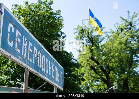 Il nome della città in ucraino Severodonetsk su un cartello stradale rotto da proiettili sullo sfondo della bandiera dell'Ucraina nel cielo, una città in Foto Stock