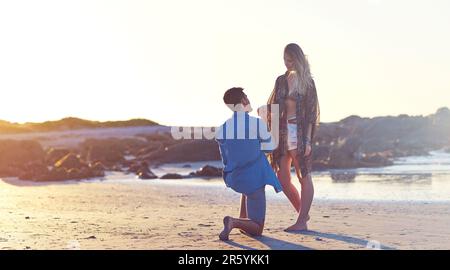 Theres qualche cosa Ive state desiderando chiederlo. un giovane che tiene la mano delle sue fidanzate sul ginocchio piegato in spiaggia. Foto Stock