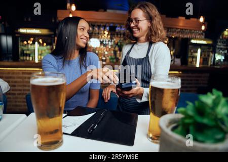 Felice giovane donna in abbigliamento casual a pagamento per bevande con tecnologia wireless accanto alla cameriera Foto Stock