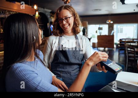 Felice giovane donna in abbigliamento casual a pagamento per la cena con la card machine da cameriera Foto Stock