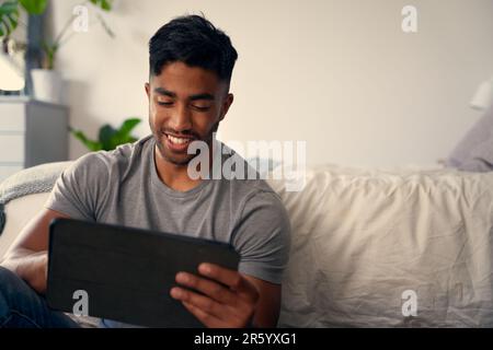 Felice giovane uomo biraciale in abbigliamento casual utilizzando il tablet digitale in camera da letto a casa Foto Stock